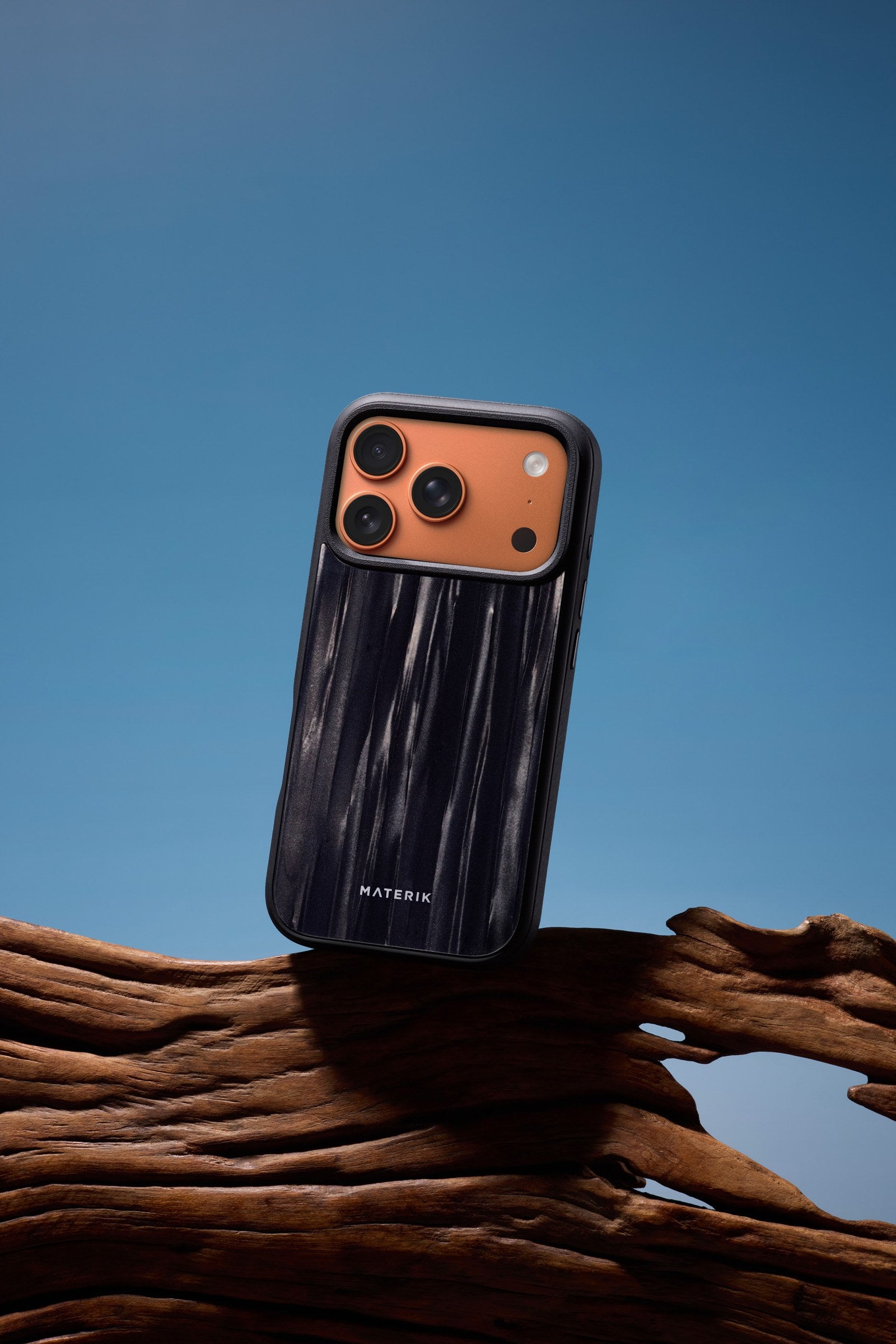 Materik PureCase for iPhone 17 Pro in black, standing on driftwood against a blue sky